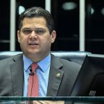 O presidente do Senado Davi Alcolumbre de paletó cinza e gravata vermelha, sentado à mesa diretora com um monitor escrito Senado Federal em primeiro plano, ilustrando a discussão sobre a PEC da escala 6x1.