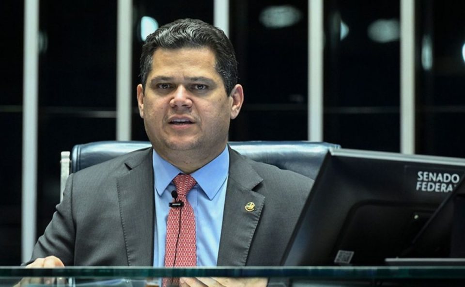 O presidente do Senado Davi Alcolumbre de paletó cinza e gravata vermelha, sentado à mesa diretora com um monitor escrito Senado Federal em primeiro plano, ilustrando a discussão sobre a PEC da escala 6x1.