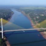 Vista aérea panorâmica da Ponte da Integração, uma ponte estaiada com cabos amarelos que cruza o Rio Paraná na fronteira entre o Brasil e o Paraguai, cercada por vegetação e áreas urbanas.