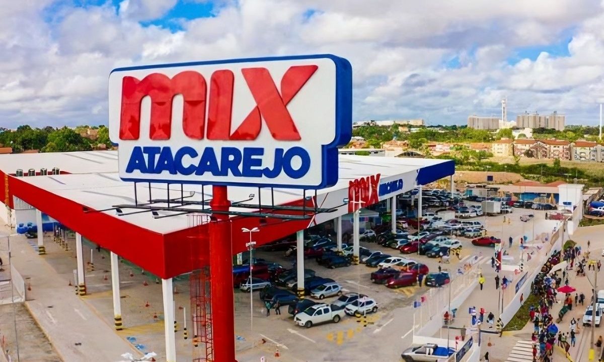 Vista aérea de uma grande loja Mix Atacarejo do Grupo Mateus, com letreiro em destaque vermelho e azul, estacionamento movimentado e rua lateral com carros, ilustrando o balanço financeiro do gigante dos supermercados.