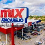 Vista aérea de uma grande loja Mix Atacarejo do Grupo Mateus, com letreiro em destaque vermelho e azul, estacionamento movimentado e rua lateral com carros, ilustrando o balanço financeiro do gigante dos supermercados.