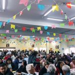 Salão paroquial lotado de pessoas sentadas em mesas e circulando, decorado com muitas bandeirinhas coloridas de festa junina penduradas no teto, durante evento festivo na Paróquia da CIC.