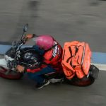Vista aérea de um entregador de aplicativo de moto em movimento em uma via pública, vestindo jaqueta vermelha, capacete rosa e carregando uma mochila de entregas laranja nas costas, destacando o público-alvo da nova linha de crédito.