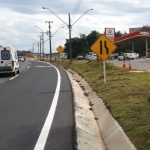 Vista de uma rodovia estadual asfaltada com uma ambulância branca em movimento, placas de sinalização amarelas à direita e um posto de combustíveis ao fundo, ilustrando o cenário de tráfego e prevenção de acidente rodovia.