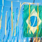 Bandeira do Brasil de plástico pendurada em fios de varal urbano, cercada por muitas fitas decorativas azuis e amarelas sob o céu azul claro, ilustrando a torcida e os cuidados contra golpes na copa do mundo.
