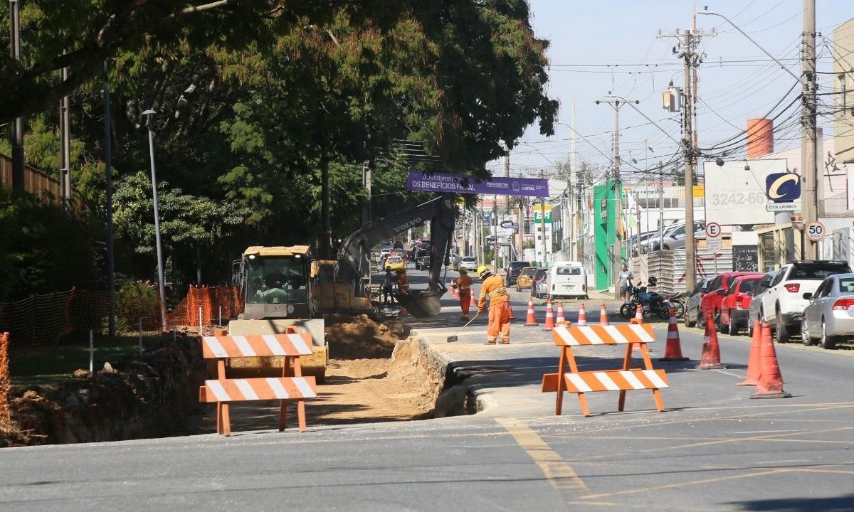 Canteiro de obras em uma avenida com operários vestindo uniformes laranjas, uma escavadeira hidráulica trabalhando na terra, cavaletes de sinalização listrados e grandes árvores ao fundo no lado esquerdo, ilustrando as obras viárias na Arthur Bernardes.