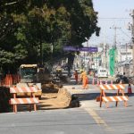 Canteiro de obras em uma avenida com operários vestindo uniformes laranjas, uma escavadeira hidráulica trabalhando na terra, cavaletes de sinalização listrados e grandes árvores ao fundo no lado esquerdo, ilustrando as obras viárias na Arthur Bernardes.