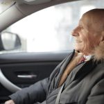 Homem idoso de perfil, sorrindo e sentado no banco de um carro de passeio enquanto usa um casaco grosso de inverno com capuz de pelos, ilustrando a matéria sobre o projeto de isenção de imposto para idosos.