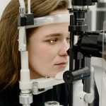 Close-up de perfil de uma mulher jovem com o rosto apoiado em um aparelho de exame oftalmológico clínico de consultório, com foco em seus olhos, ilustrando a avaliação para concessão de benefício de carro com desconto.