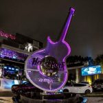 Fachada noturna do restaurante Hard Rock Cafe em Curitiba com uma enorme escultura iluminada em formato de guitarra com luzes neon roxas na entrada principal, onde ocorrerá o encontro promovido pelo Terceiro Setor Curitiba.