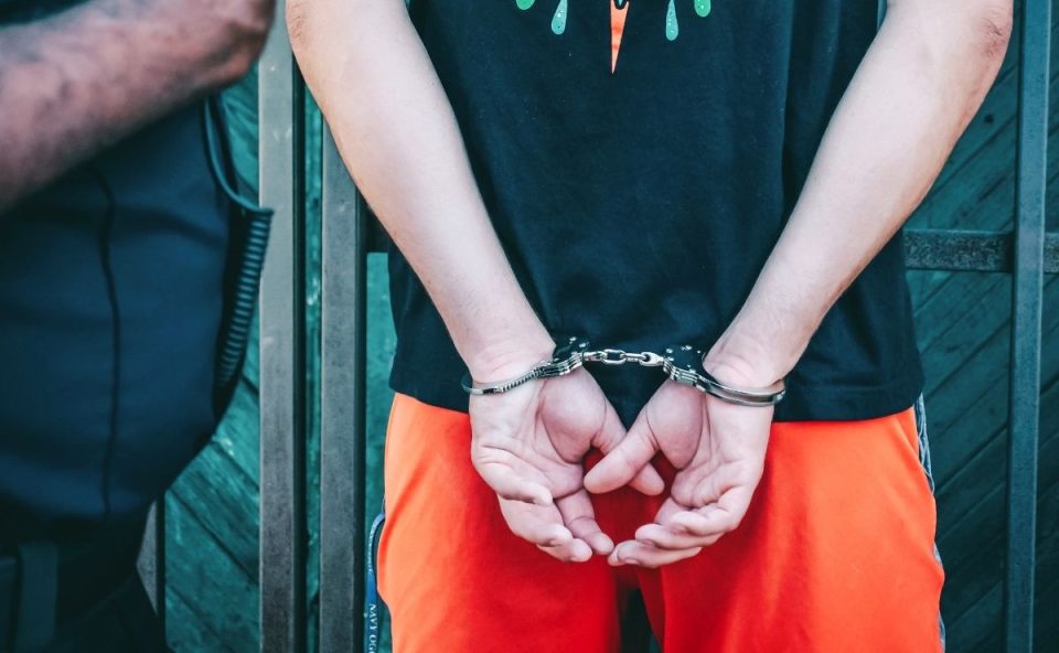Close-up das costas de um jovem vestindo camiseta preta e bermuda laranja, com as mãos algemadas para trás ao lado de um policial, ilustrando o debate sobre o projeto que reduz maioridade penal.