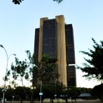 Vista externa em ângulo baixo do Edifício-Sede do Banco Central do Brasil em Brasília, um arranha-céu de arquitetura brutalista com janelas escuras, cercado por árvores e postes de iluminação sob o céu claro, ilustrando o debate sobre a autonomia do Banco Central.