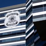 Fachada moderna e espelhada de um edifício com listras horizontais em tons de cinza, branco e preto, exibindo em destaque o brasão oficial da Polícia Federal, ilustrando a fiscalização do setor de Segurança Privada.