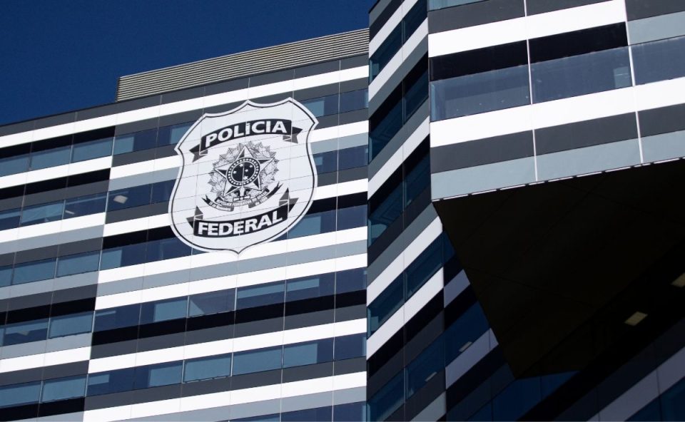 Fachada moderna e espelhada de um edifício com listras horizontais em tons de cinza, branco e preto, exibindo em destaque o brasão oficial da Polícia Federal, ilustrando a fiscalização do setor de Segurança Privada.