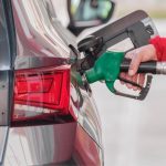 Close-up da mão de um frentista usando casaco vermelho e segurando um bico de bomba de combustível verde inserido no tanque de um carro cinza, ilustrando o abastecimento de veículos com gasolina sem imposto.