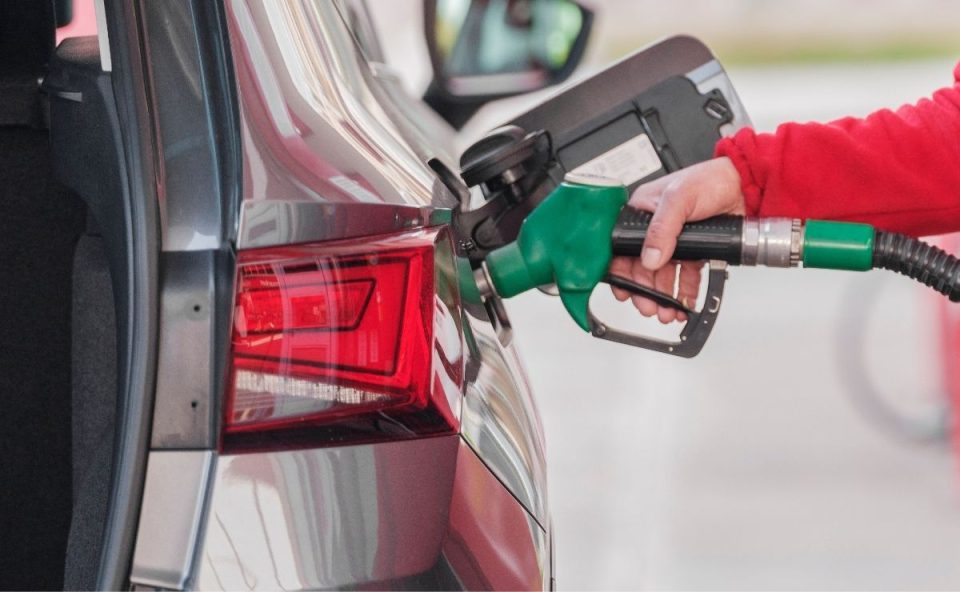 Close-up da mão de um frentista usando casaco vermelho e segurando um bico de bomba de combustível verde inserido no tanque de um carro cinza, ilustrando o abastecimento de veículos com gasolina sem imposto.