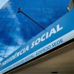 Vista em ângulo diagonal de uma grande placa azul com letras brancas onde se lê "Previdência Social - Instituto Nacional do Seguro Social - Edifício Sede", fixada na entrada de um prédio moderno com fachada espelhada que reflete o céu azul, ilustrando as ações judiciais de aposentados contra o INSS.