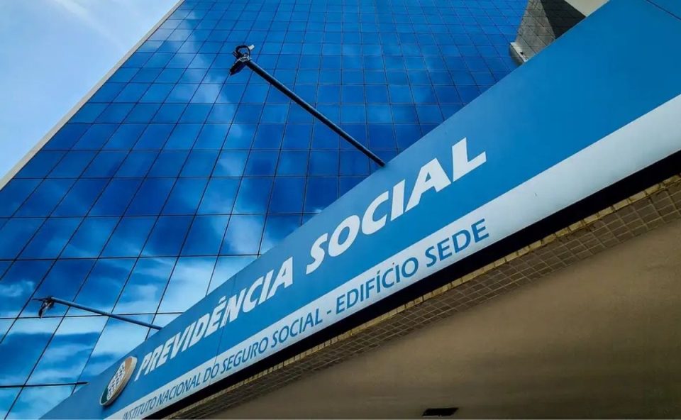 Vista em ângulo diagonal de uma grande placa azul com letras brancas onde se lê "Previdência Social - Instituto Nacional do Seguro Social - Edifício Sede", fixada na entrada de um prédio moderno com fachada espelhada que reflete o céu azul, ilustrando as ações judiciais de aposentados contra o INSS.