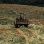 Vista traseira de um trator agrícola vermelho guiado por um operador, transitando por uma estrada de terra em meio a um campo verdejante e uma lavoura em terreno inclinado, ilustrando o trabalho diário e os benefícios econômicos para produtores rurais.