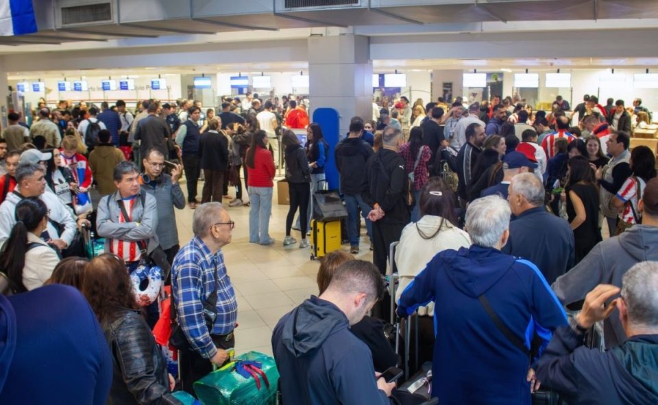 Vista interna do saguão de embarque lotado de passageiros com malas em filas de check-in, destacando pessoas de costas e de perfil esperando atendimento no Aeroporto de Assunção durante o fluxo da Copa do Mundo.