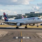 Avião da Latam Airlines pousando na pista do aeroporto, com o terminal e a torre de controle visíveis ao fundo. Há outros aviões e veículos estacionados. A imagem acompanha a notícia sobre o aumento de voos direto de Maringá para São Paulo, fortalecendo a conectividade aérea no Norte do Paraná.