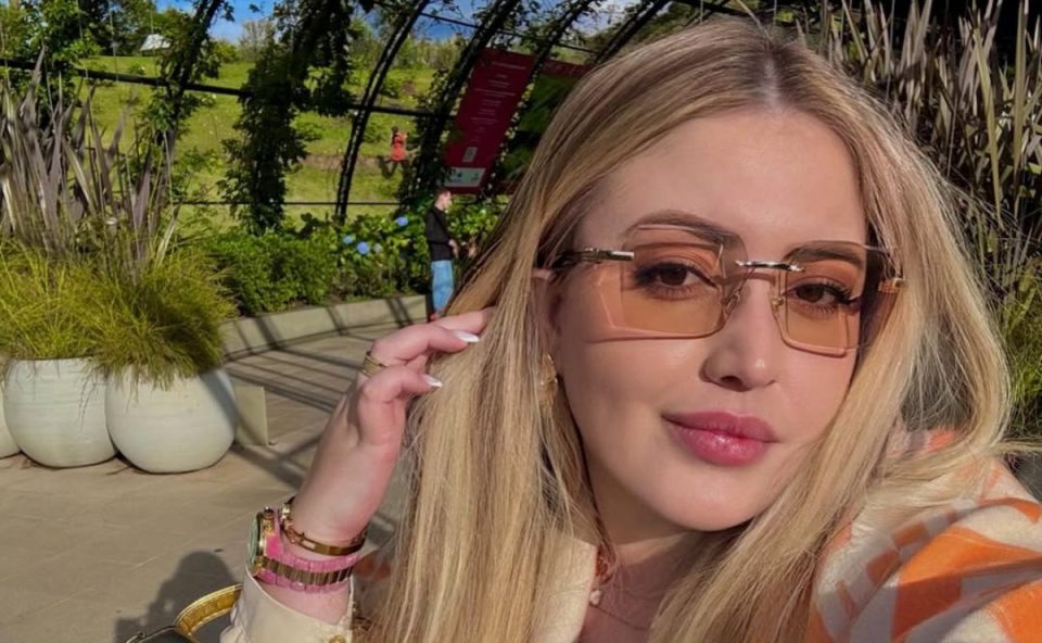 foto mostra allana brittes. ela é uma mulher loira e branca . está com óculos transparente, batom rosa e mão no cabelo.