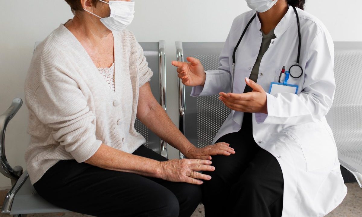 Profissional da área da saúde conversa com paciente em unidade hospitalar durante atendimento presencial, ambas utilizando máscaras de proteção.