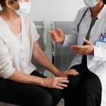 Profissional da área da saúde conversa com paciente em unidade hospitalar durante atendimento presencial, ambas utilizando máscaras de proteção.