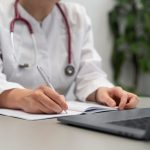 foto ilustrativa sobre a notícia do atestado falso na grande curitiba. foto mostra uma médica com estetoscopoio na frente de um notebook.