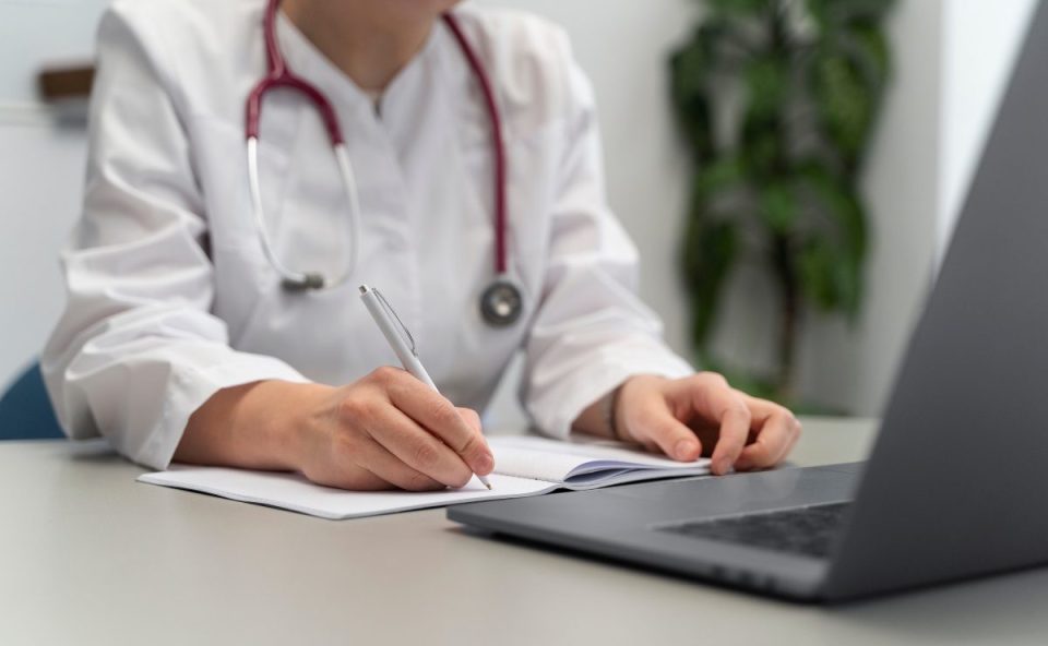 foto ilustrativa sobre a notícia do atestado falso na grande curitiba. foto mostra uma médica com estetoscopoio na frente de um notebook.