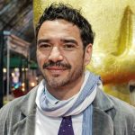 A imagem mostra o ator Caio Blat que contou como foi o incidente que apnhou de cinta no inicio da carreira ele está sorrindo e com um cachecou no pescoço