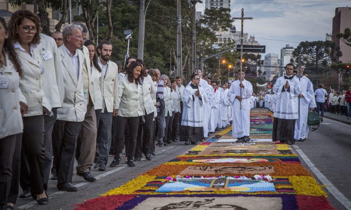 Fiéis participam da procissão de Corpus Christi em Curitiba, caminhando ao lado dos tradicionais tapetes coloridos confeccionados nas ruas da cidade.