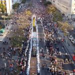 Vista aérea da celebração de Corpus Christi em Curitiba, com milhares de fiéis acompanhando a procissão ao longo da Avenida Cândido de Abreu, onde um extenso tapete religioso colorido ocupa o centro da via.