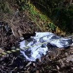 Imagem mostra a área isolada onde a criança foi encontrada morta em uma estrutura de esgoto. Um material cobre o corpo da vítima. Ao redor, há galhos secos, entulhos e vegetação. A cena ocorre durante a noite, com pessoas acompanhando a ocorrência próximas ao local.