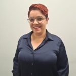 Cíntia Rocha, editora da Banda B, com camisa azul escura e em frente a uma parede cinza