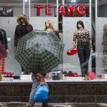Vitrine de loja decorada para o Dia dos Namorados, com manequins, balões vermelhos e a frase "Te Amo", enquanto uma pessoa caminha na chuva carregando um guarda-chuva.
