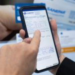 Pessoa segura um smartphone exibindo uma fatura da Sanepar na tela, enquanto aponta para informações da conta para renegociar dívidas Ao fundo, aparecem documentos da companhia desfocados.