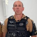 Fotografia mostra um policial militar do Paraná morto por dupla durante confronto. O homem branco e careca aparece fardado, em pé, com postura séria e os braços para trás, diante de um fundo branco.
