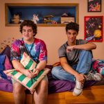 Foto com os atores Miguel Lallo e Diego Lira, do filme "Quinze Dias". Os dois são adolescentes e aparecem sentados à uma cama, em um quarto colorido.