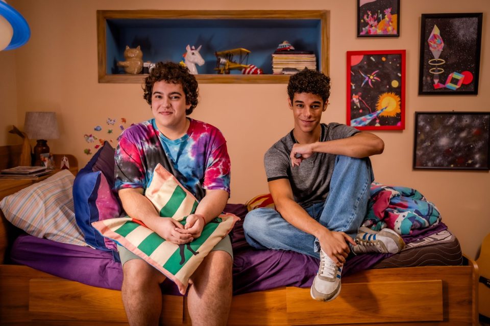 Foto com os atores Miguel Lallo e Diego Lira, do filme "Quinze Dias". Os dois são adolescentes e aparecem sentados à uma cama, em um quarto colorido.