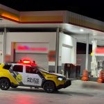 Foto mostra viatura da Polícia Militar parada no pátio de um posto de gasolina durante a noite e o corpo de um homem próximo ao carro. Homem morreu após agredir clientes, passar mal e cair, segundo a PM.