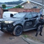Foto tirada durante a manhã mostra policial fardado e armado ao lado de viatura preta da Polícia Militar. Ele está em endereço onde suspeito de fabricar armas para facção morreu em confronto