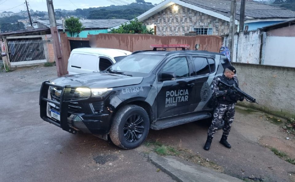 Foto tirada durante a manhã mostra policial fardado e armado ao lado de viatura preta da Polícia Militar. Ele está em endereço onde suspeito de fabricar armas para facção morreu em confronto
