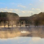Neblina sobre lago durante manhã fria em parque de Curitiba, com passarela de madeira e pessoas caminhando ao nascer do sol.