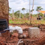 A imagem mostra um registro de água instalado em uma área de terra vermelha. Ao fundo, desfocados, aparecem trabalhadores realizando serviços, além de um carro, uma motocicleta e uma tenda. O cenário é externo, durante o dia, sob céu nublado
