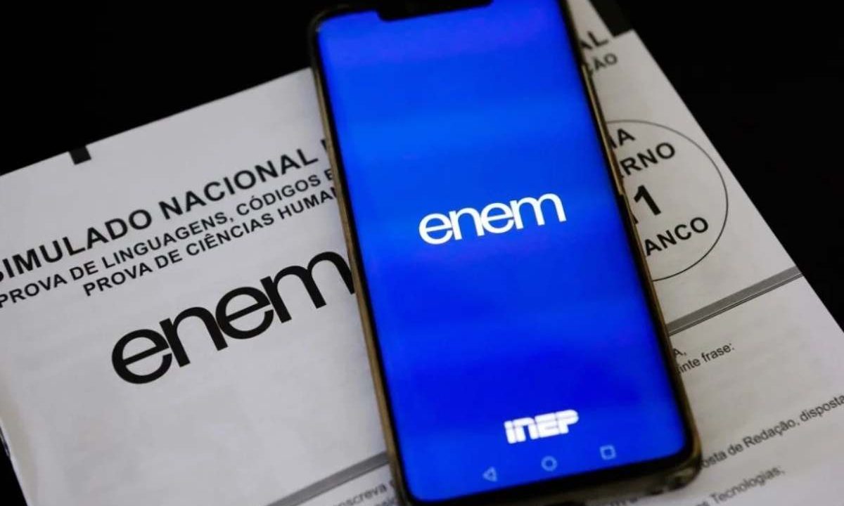 Foto da tela de um celular entrando na Página do Participante. Ao fundo, em baixo do celular, um prova do Enem. Imagem ilustrativa para matéria: Enem 2026 tem novas condições para atendimento especializado; veja o que muda