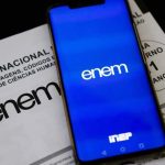 Foto da tela de um celular entrando na Página do Participante. Ao fundo, em baixo do celular, um prova do Enem. Imagem ilustrativa para matéria: Enem 2026 tem novas condições para atendimento especializado; veja o que muda