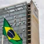 Prejuízo dos Correios chega a R$ 3,16 bilhões no primeiro trimestre de 2026. A imagem mostra o prédio sede dos Correios ao lado de uma bandeira do Brasil.