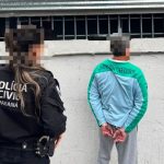 Policiais civis do Paraná acompanham homem algemado durante cumprimento de mandado de prisão em Curitiba. Suspeito aparece de costas, em frente a um muro, com o rosto desfocado.