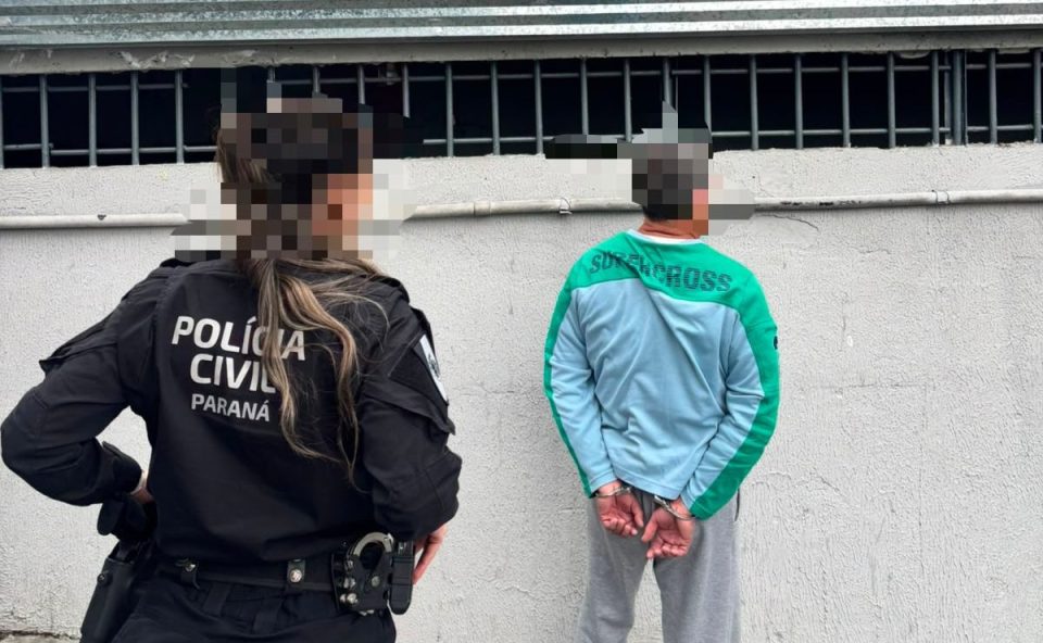 Policiais civis do Paraná acompanham homem algemado durante cumprimento de mandado de prisão em Curitiba. Suspeito aparece de costas, em frente a um muro, com o rosto desfocado.